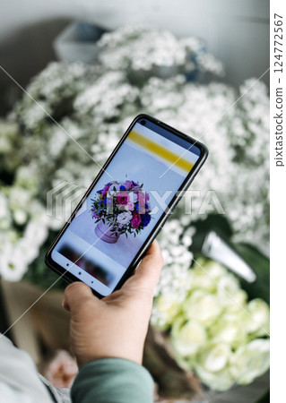 Person browsing a flower bouquet on a smartphone screen surrounded by real white blossoms. Flower subscription, floral e-commerce, gift delivery services, digital floristry 124772567