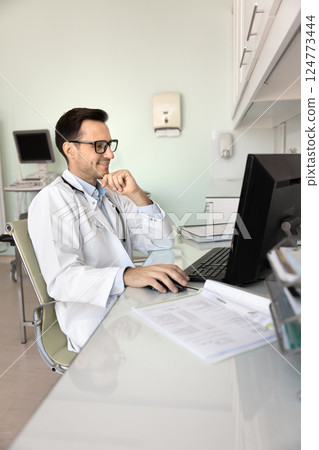 Satisfied male doctor sitting at desk, working on computer Satisfied male doctor sitting at desk, working on computer 124773444
