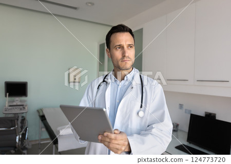 Thoughtful professional male GP doctor in white coat using tablet Thoughtful professional male GP doctor in white coat using tablet 124773720