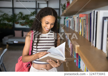 Excited teen girl student standing by bookcase turning book pages Excited teen girl student standing by bookcase turning book pages 124773841