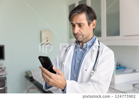 Physician in coat using smartphone in consulting room 124773902