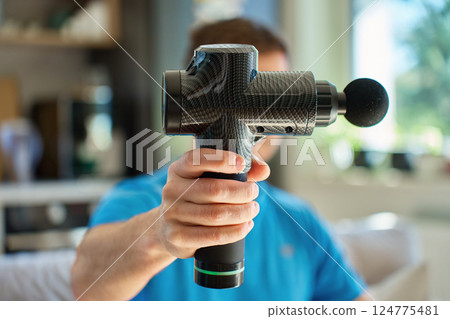 Close-up of man in blue shirt holding black percussion massage gun in living room. Concept of sports recovery, self-care, and muscle treatment tools Close-up of man in blue shirt holding black percussion massage gun in living room. Concept of sports recovery, self-care, and muscle treatment tools 124775481