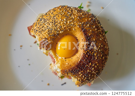 Close-up of bitten bagel with egg, ham, cheese and fresh greens. Half-eaten breakfast sandwich on plate, top view 124775512