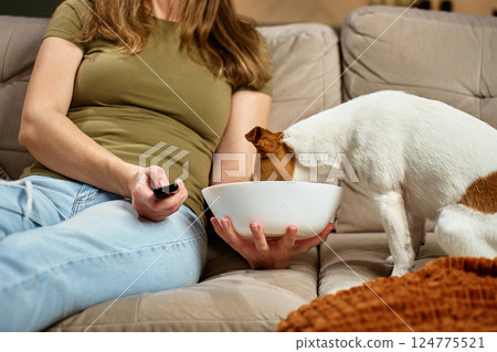 Woman relaxing on sofa with remote control while dog steals food. Pet dog eat food from bowl while owner focuses on TV. Home life and pet behavior concept 124775521