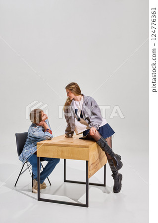 Two students interact near wooden desk against plain background. Boy sits on black chair, looking up at girl, who leans on desk. Two students interact near wooden desk against plain background. Boy sits on black chair, looking up at girl, who leans on desk. 124776361