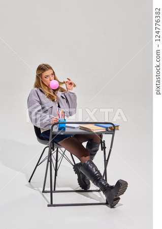 Blonde girl in grey jacket and black boots blows pink bubblegum bubble while sitting at desk with books, notebook, and cup of colorful pens. 124776382