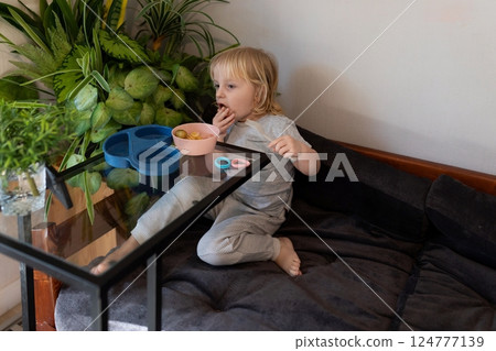 Child enjoys snack at home sitting on sofa with plants nearby in a cozy indoor setting 124777139