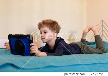 Young child enjoys playing games on a tablet while lounging on a cozy bed in a bright room Young child enjoys playing games on a tablet while lounging on a cozy bed in a bright room 124777546