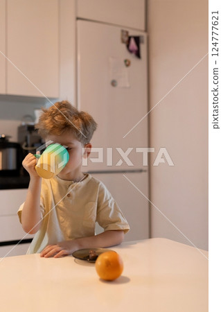 Young boy enjoying a warm drink at a kitchen table in the morning with a fresh orange nearby Young boy enjoying a warm drink at a kitchen table in the morning with a fresh orange nearby 124777621