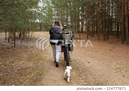 Woman walks with child on bicycle in forest while dog runs alongside them during a sunny afternoon Woman walks with child on bicycle in forest while dog runs alongside them during a sunny afternoon 124777671