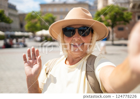 Senior woman taking a selfie, waving hand in a city square, enjoying vacation. 124778226