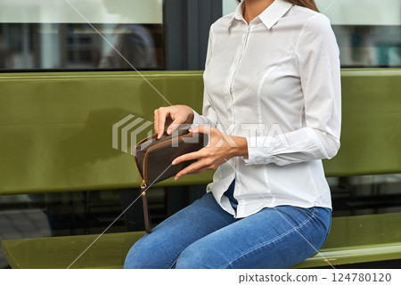 Woman in white shirt, jeans, holding leather wallet, sitting on green bench, outdoor, style, casual, urban lifestyle, fashion concept Woman in white shirt, jeans, holding leather wallet, sitting on green bench, outdoor, style, casual, urban lifestyle, fashion concept 124780120