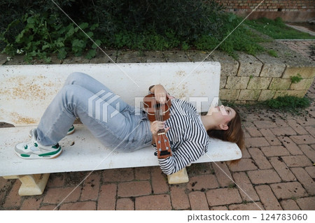 Artist Girl Plays Musical Instrument Ukulele Lying On Bench In Park Artist Girl Plays Musical Instrument Ukulele Lying On Bench In Park 124783060