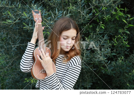 Artist Girl Plays Ukulele Musical Instrument Outside In Park Near A Tree 124783077