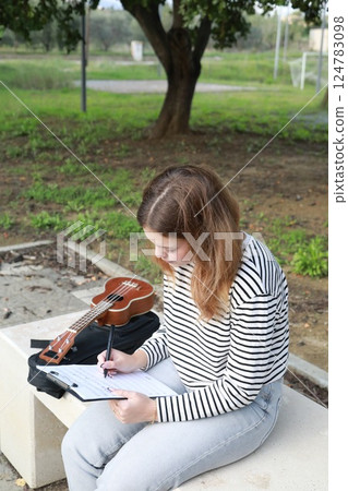 Girl Writes Notes On A Sheet Music To Play Them With The Ukulele 124783098