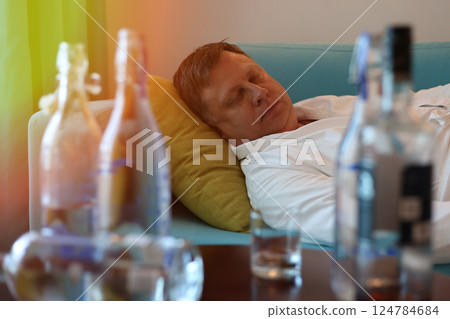 A middle aged man rests on a sofa near a table filled with bottles of alcohol, tired and sore after drinking too much 124784684