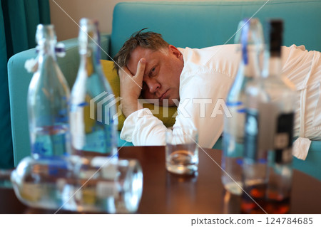 A middle aged man rests on a sofa near a table filled with bottles of alcohol, tired and sore after drinking too much 124784685