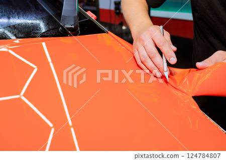 Skilled technician carefully cuts applies vibrant orange vinyl wrap to car surface at an auto detailing shop. Skilled technician carefully cuts applies vibrant orange vinyl wrap to car surface at an auto detailing shop. 124784807