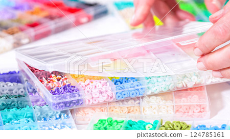 Woman hands gracefully poised over a collection of beads, sorted by color in transparent organizers. The array of beads spans a vibrant spectrum, from deep purples to bright oranges, meticulously 124785161