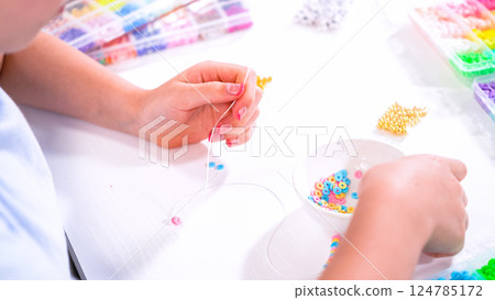 Delicate fingers of a young girl navigate through a treasure trove of bright, multicolored beads, each compartment revealing a new hue to choose from. She's immersed in the joyful task of stringing 124785172