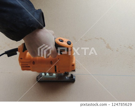 hand presses orange jigsaw and moves along marking line, cutting sheet of pressed wood into pieces, cutting sheet of plywood lengthwise with jigsaw along vertical marking line 124785629