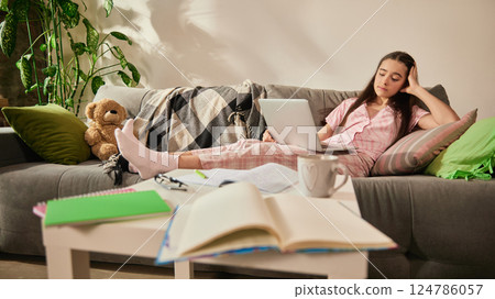Middle school student, girl lying on couch in pink pajamas with tired expression, holding head in hand, struggling with long online learning session. Middle school student, girl lying on couch in pink pajamas with tired expression, holding head in hand, struggling with long online learning session. 124786057
