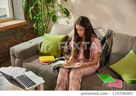 Concentrated teen girl, middle school student in pajamas studying with open notebooks and laptop, writing school notes on sofa 124786065