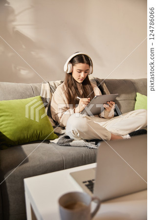 Focused teen girl sitting cross-legged on sofa with headphones, writing on digital tablet during remote learning session in modern room interior Focused teen girl sitting cross-legged on sofa with headphones, writing on digital tablet during remote learning session in modern room interior 124786066