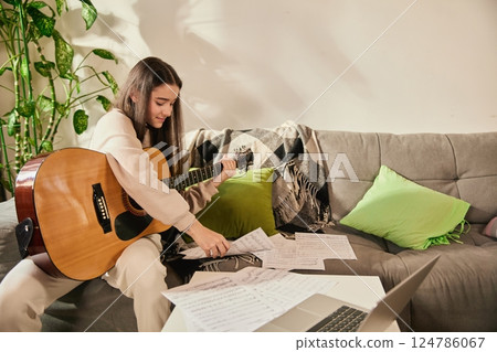 Smiling teen girl on couch at home sitting with guitar and learning notes, focusing on learning music and rhythm structure Smiling teen girl on couch at home sitting with guitar and learning notes, focusing on learning music and rhythm structure 124786067