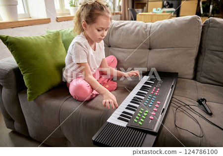 Focused child sitting cross-legged on couch at home and playing electronic keyboard during online music activity. Focused child sitting cross-legged on couch at home and playing electronic keyboard during online music activity. 124786100