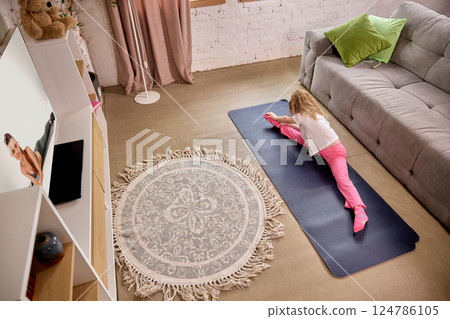 Girl doing deep stretch at home while watching virtual fitness lesson on TV in bright cozy room. Girl doing deep stretch at home while watching virtual fitness lesson on TV in bright cozy room. 124786105