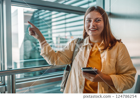 Happy young woman pointing and smiling at the airport terminal while holding a smartphone. 124786254
