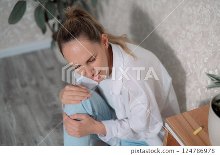 Young woman sitting discomfort in her injury knee pain while adjusting her posture in a cozy indoor setting 124786978