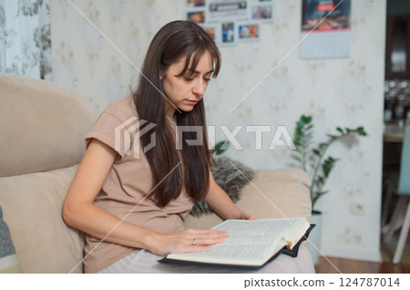 Millennial woman reading book bible while relaxing on comfortable sofa, immersed in quiet moment of solitude at home 124787014