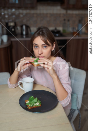 Young woman eating avocado toast healthy food, drinking herbal tea near bright kitchen window 124787019