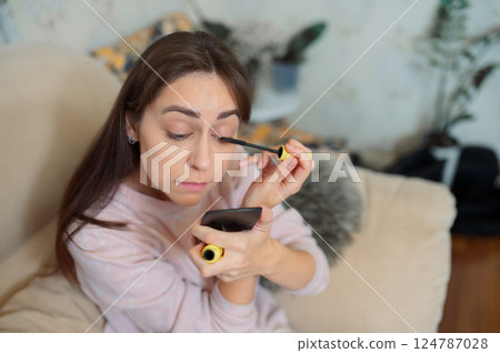 Young woman applying mascara, gazing into handheld mirror while relaxing on living room sofa with soft background lighting morning routine Young woman applying mascara, gazing into handheld mirror while relaxing on living room sofa with soft background lighting morning routine 124787028