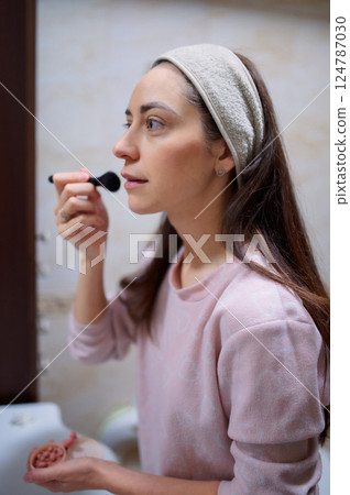 Woman 20s in bathrobe applying makeup, brushing cosmetics while standing before bathroom mirror morning routine Woman 20s in bathrobe applying makeup, brushing cosmetics while standing before bathroom mirror morning routine 124787030