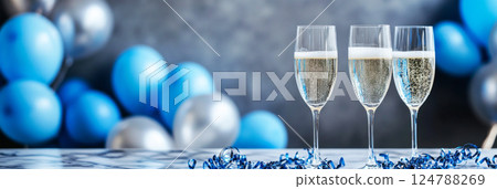A modern, festive party scene with bubbly wine in elegant crystal glasses, blue balloons, and metallic streamers, setting a luxurious and joyful mood for a chic celebration, banner 124788269
