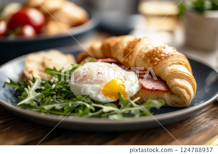 A rustic croissant filled with poached egg, ham, and arugula, illuminated by soft natural window light. The light highlights the layers and creates gentle shadows, adding depth and warmth 124788937