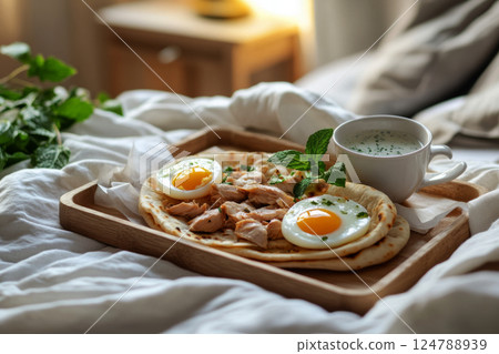 A dark wooden tray with pita filled with chicken, boiled egg, and yogurt dressing, paired with mint tea, placed on white linen bed in a cozy, warm modern bedroom A dark wooden tray with pita filled with chicken, boiled egg, and yogurt dressing, paired with mint tea, placed on white linen bed in a cozy, warm modern bedroom 124788939