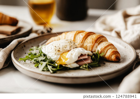 A flaky croissant filled with a poached egg, ham, and fresh arugula leaves, set in a cozy rustic scene. Soft, diffused lighting enhances the rich textures and natural warmth A flaky croissant filled with a poached egg, ham, and fresh arugula leaves, set in a cozy rustic scene. Soft, diffused lighting enhances the rich textures and natural warmth 124788941
