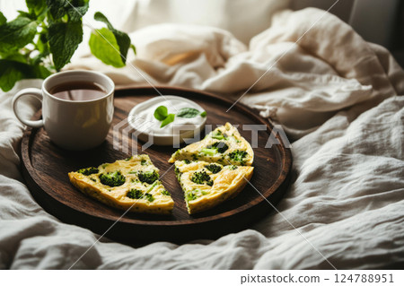 A cozy morning setup featuring frittata triangles with broccoli, goat cheese, and Parmesan, paired with a fragrant cup of tea, placed on a wooden tray, surrounded by soft linen in a modern bedroom A cozy morning setup featuring frittata triangles with broccoli, goat cheese, and Parmesan, paired with a fragrant cup of tea, placed on a wooden tray, surrounded by soft linen in a modern bedroom 124788951
