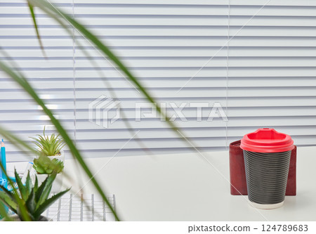 Office desk with white blinds in the background. Gray and red coffee cup sits on the desk, along with a green plant and a wire mesh organizer. Office desk with white blinds in the background. Gray and red coffee cup sits on the desk, along with a green plant and a wire mesh organizer. 124789683