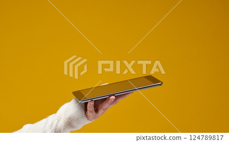 Hand wearing a white sweater holds a silver tablet against a solid yellow background. The tablet screen is blank and facing upwards, copy space 124789817