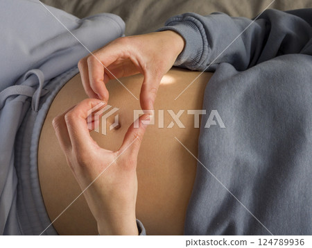 Expectant mother reclining, creating heart gesture over rounded abdomen, radiating maternal connection and tender anticipation of newborn arrival 124789936