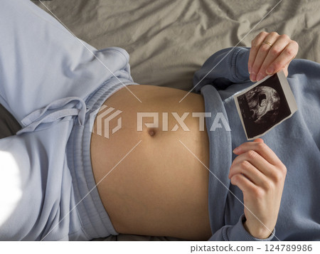 Expectant mother resting comfortably in bed, wearing soft blue loungewear, tenderly cradling ultrasound image against pregnant belly, embracing maternal joy and anticipation 124789986