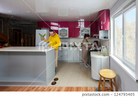 Husband holds tulips in kitchen with red cabinets, wife cooks near stove, wide angle lens captures bright light and domestic activity near island. 124793405