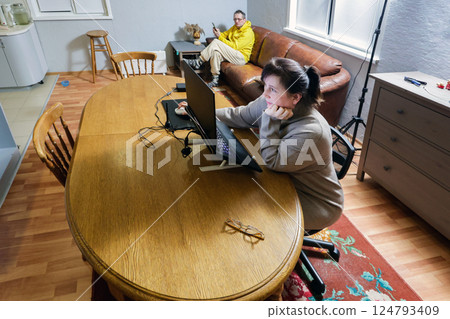 Wife works on laptop at wooden table, glasses sit on table, husband relaxes on couch, wide angle shot of living space. 124793409