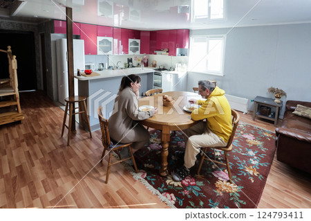 Two people are having meal together in kitchen with pink cabinets, sitting at wooden table, basket of bread visible, floral rug on floor. Two people are having meal together in kitchen with pink cabinets, sitting at wooden table, basket of bread visible, floral rug on floor. 124793411