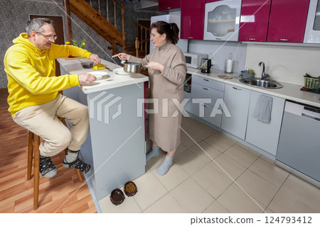 Wife serves food at kitchen island while husband sits on bar stool nearby, pet bowls sit on floor in spacious kitchen with wooden accents. Wife serves food at kitchen island while husband sits on bar stool nearby, pet bowls sit on floor in spacious kitchen with wooden accents. 124793412